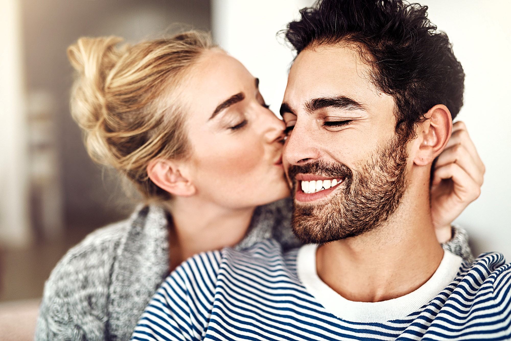 A woman with blonde hair is gently kissing a smiling, bearded man on the cheek. They are indoors, and the man is wearing a striped shirt while the woman wears a cozy sweater. The scene conveys warmth and affection.