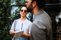 A woman wearing sunglasses and holding a notepad stands outside near greenery. She looks attentively forward, while a man in focus in the foreground appears in profile, slightly obscuring the view.