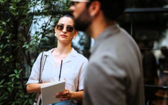 A woman wearing sunglasses and holding a notepad stands outside near greenery. She looks attentively forward, while a man in focus in the foreground appears in profile, slightly obscuring the view.