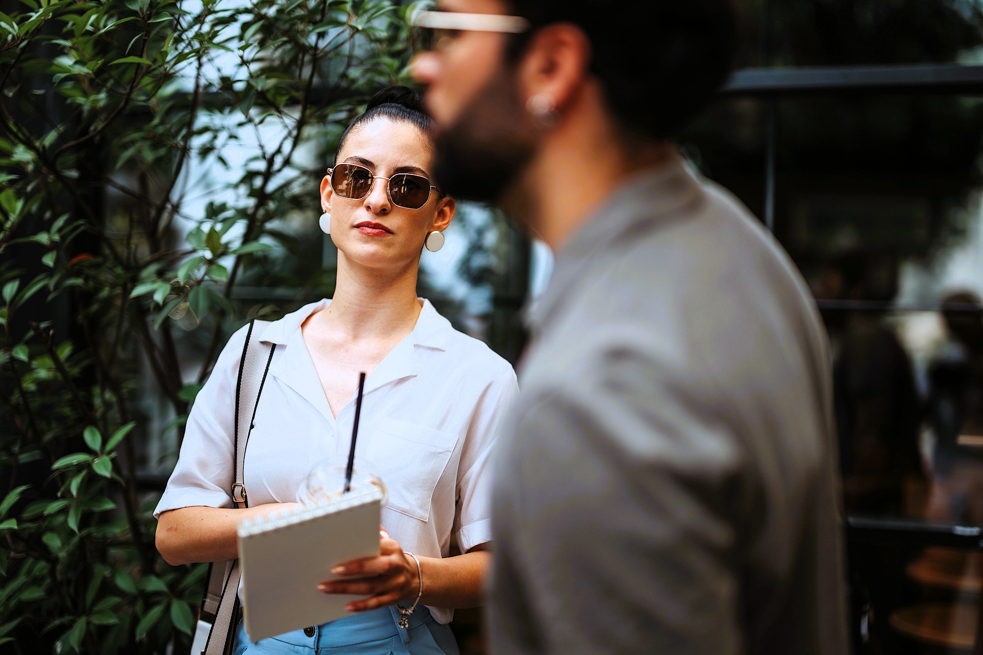 A woman wearing sunglasses and holding a notepad stands outside near greenery. She looks attentively forward, while a man in focus in the foreground appears in profile, slightly obscuring the view.