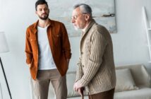 A young man in a brown jacket stands behind an older man in a beige sweater using a walking cane. They are indoors with a neutral-toned background and a blurred painting.