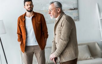 A young man in a brown jacket stands behind an older man in a beige sweater using a walking cane. They are indoors with a neutral-toned background and a blurred painting.