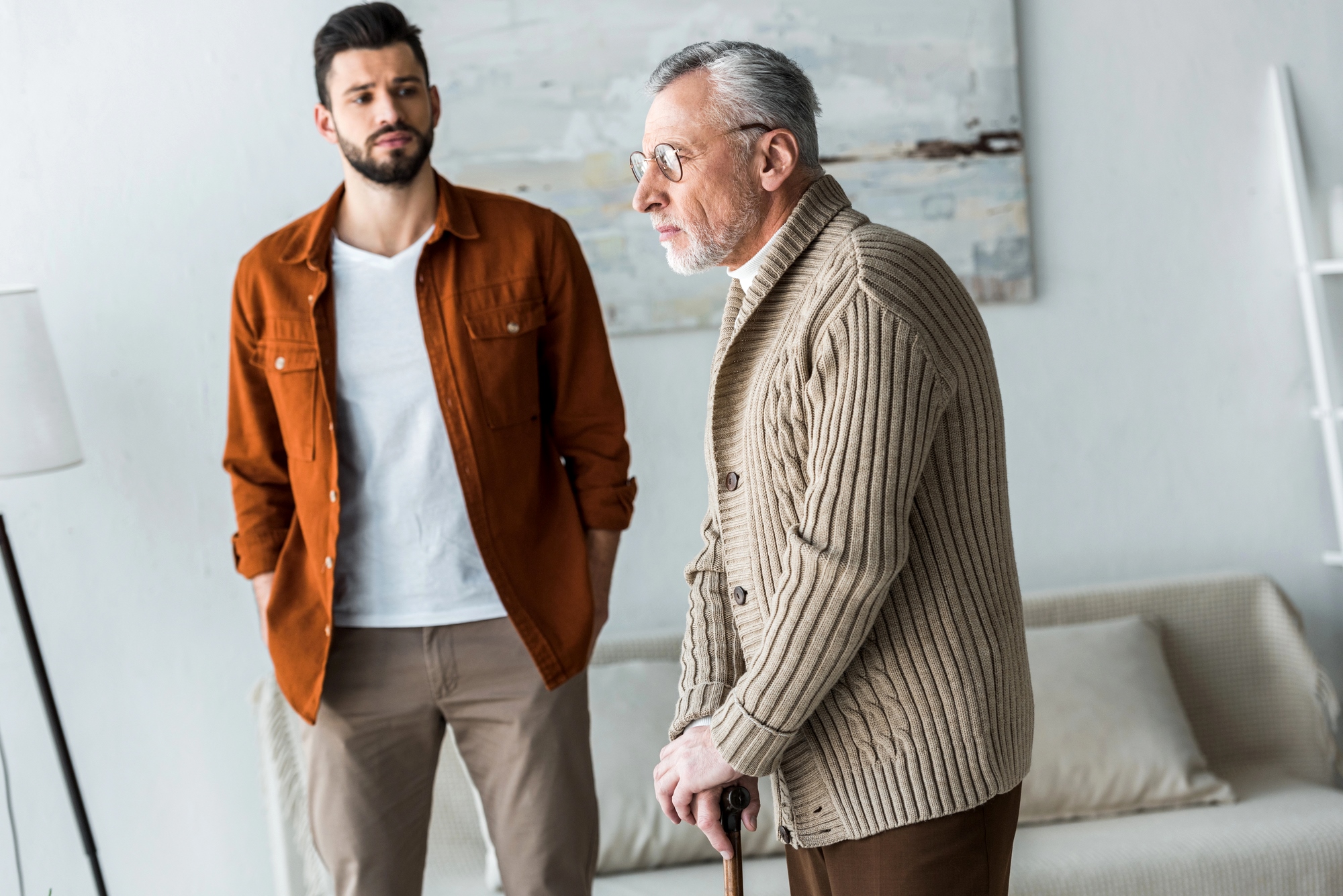 A young man in a brown jacket stands behind an older man in a beige sweater using a walking cane. They are indoors with a neutral-toned background and a blurred painting.