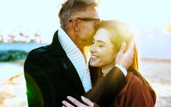 A couple embraces outdoors, smiling as sunlight beams around them. The man is wearing sunglasses and a scarf, while the woman looks content. The background features a blurred beach scene with vibrant lighting effects.