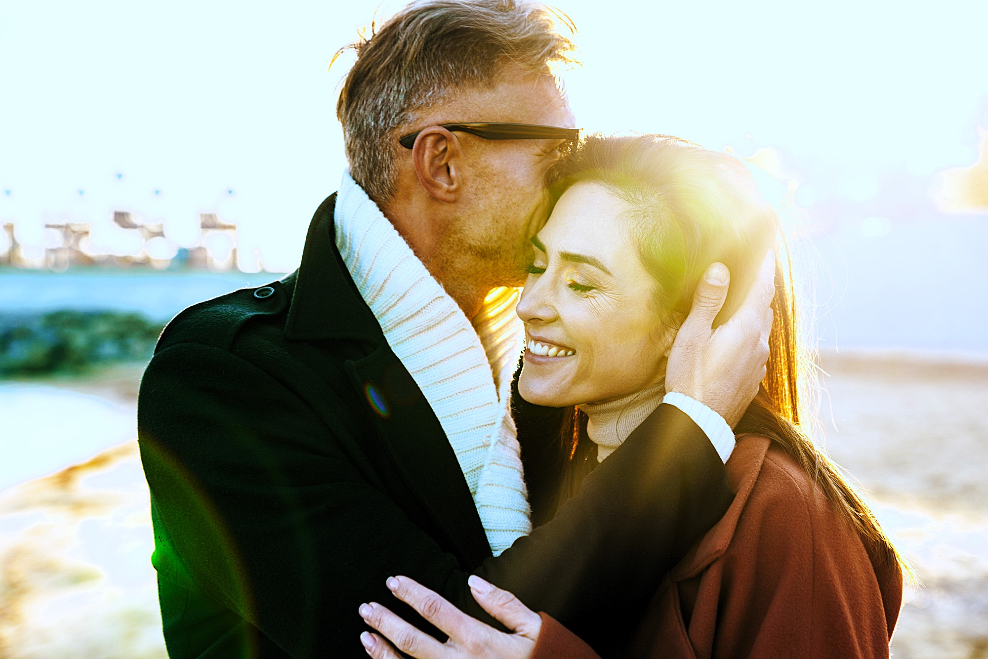 A couple embraces outdoors, smiling as sunlight beams around them. The man is wearing sunglasses and a scarf, while the woman looks content. The background features a blurred beach scene with vibrant lighting effects.