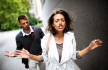 A woman in a white coat appears frustrated, walking away with her hands raised. A man in a gray sweater follows her, gesturing with one hand. They are outdoors, with a blurred background of trees and buildings.