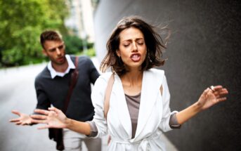 A woman in a white coat appears frustrated, walking away with her hands raised. A man in a gray sweater follows her, gesturing with one hand. They are outdoors, with a blurred background of trees and buildings.