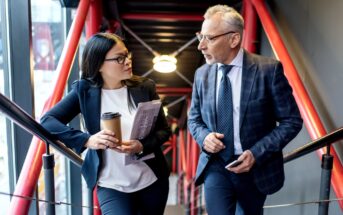 A woman and a man in business attire are walking and talking on a modern stairway with red railings. The woman holds a coffee cup and a folder, while the man is holding a smartphone. They appear engaged in a serious discussion.