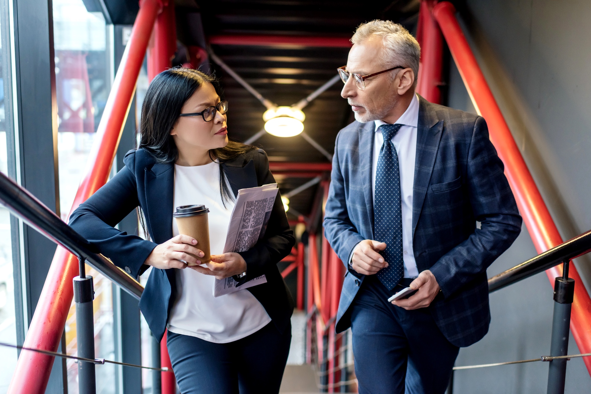 A woman and a man in business attire are walking and talking on a modern stairway with red railings. The woman holds a coffee cup and a folder, while the man is holding a smartphone. They appear engaged in a serious discussion.