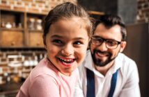 A young girl with a joyful expression looks to the side, while a man wearing glasses and smiling sits behind her. They are in a cozy kitchen setting with brick walls and wooden shelves.