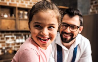 A young girl with a joyful expression looks to the side, while a man wearing glasses and smiling sits behind her. They are in a cozy kitchen setting with brick walls and wooden shelves.