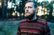 A man with a beard stands in a forest, wearing a colorful patterned sweater. The background shows trees with soft sunlight filtering through, creating a serene atmosphere.