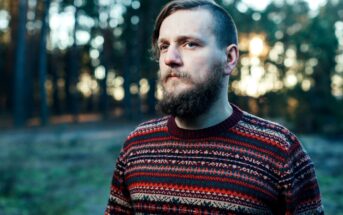 A man with a beard stands in a forest, wearing a colorful patterned sweater. The background shows trees with soft sunlight filtering through, creating a serene atmosphere.