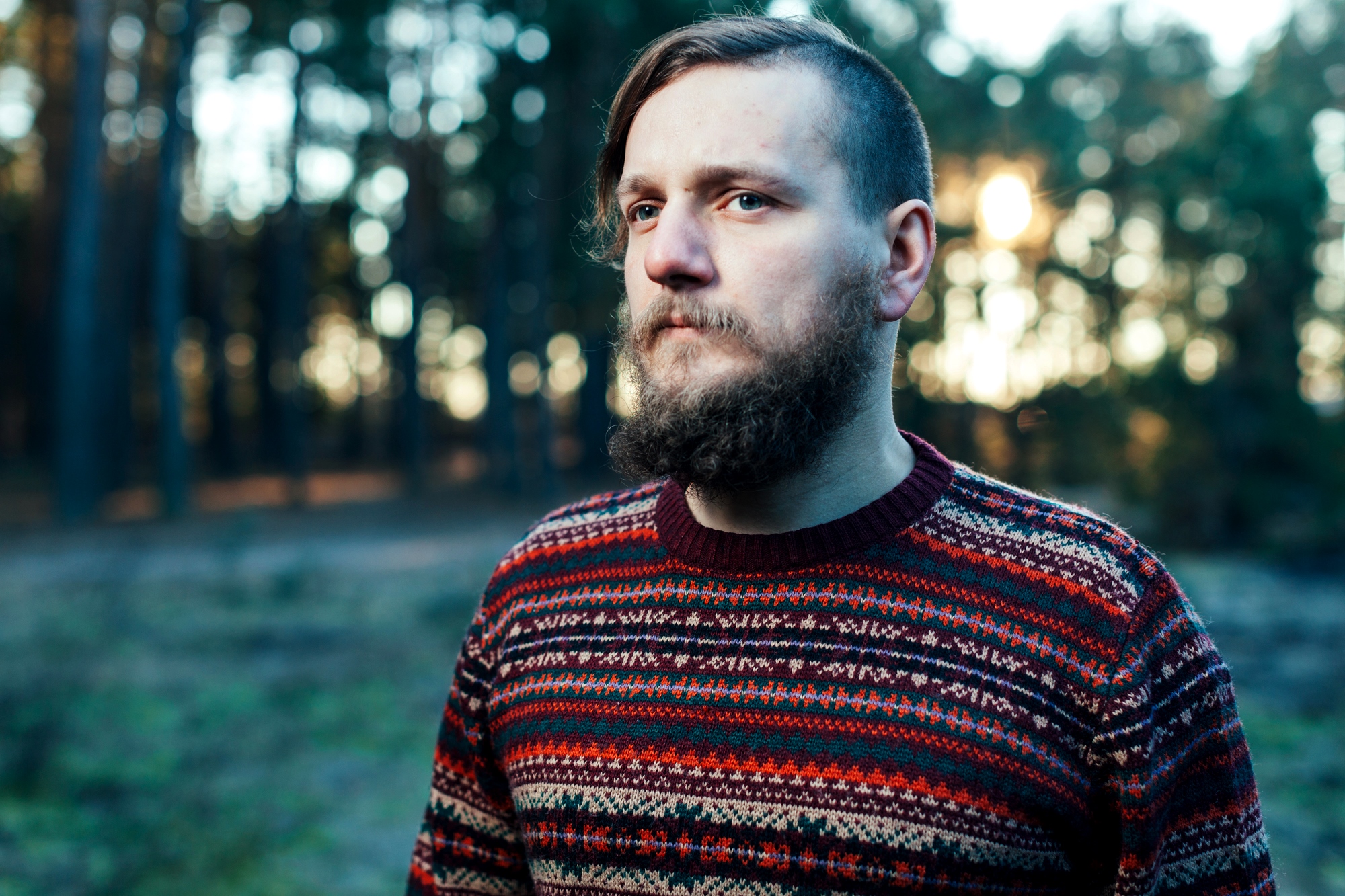 A man with a beard stands in a forest, wearing a colorful patterned sweater. The background shows trees with soft sunlight filtering through, creating a serene atmosphere.