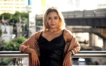 A woman with blonde hair leans against a railing, wearing a black dress and a light jacket. The background shows a cityscape with trees, buildings, and an overpass. The lighting is bright and natural.