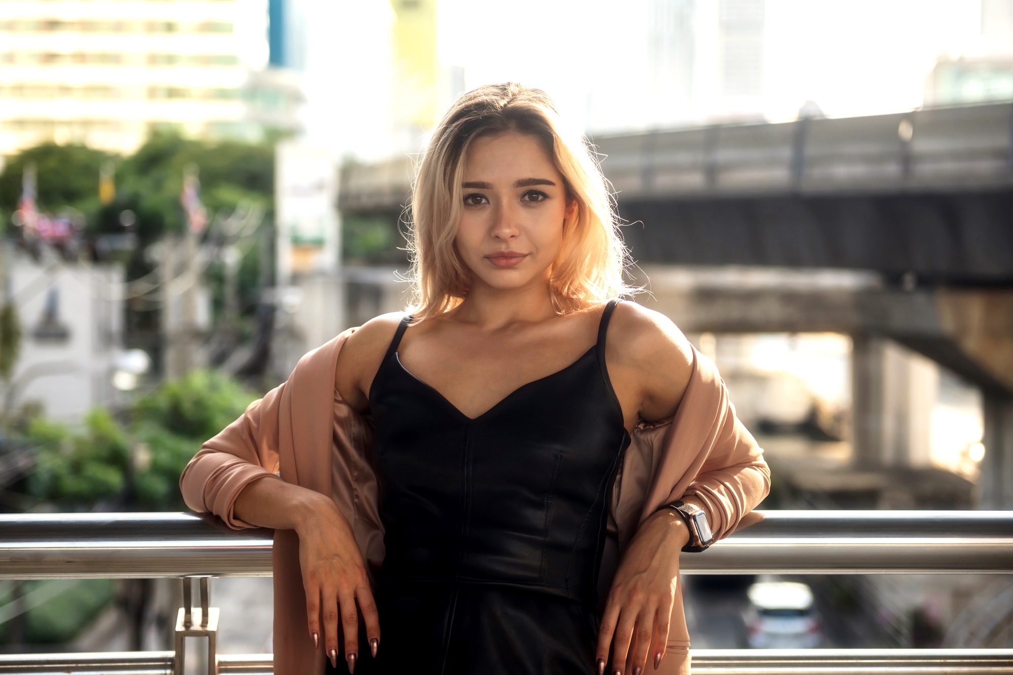 A woman with blonde hair leans against a railing, wearing a black dress and a light jacket. The background shows a cityscape with trees, buildings, and an overpass. The lighting is bright and natural.