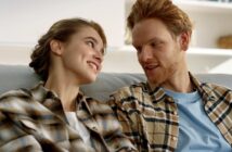 A couple wearing plaid shirts sits on a couch, smiling and looking into each other's eyes. They appear relaxed and content in a cozy indoor setting.