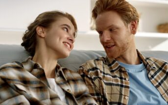 A couple wearing plaid shirts sits on a couch, smiling and looking into each other's eyes. They appear relaxed and content in a cozy indoor setting.