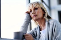 A woman with blonde hair and a thoughtful expression leans her head on her hand while looking out of a window. She is wearing a gray sweater and holding a mug, sitting in a brightly lit room.