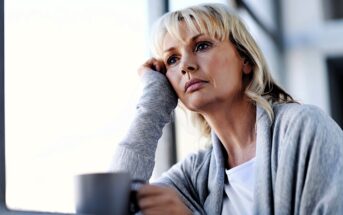 A woman with blonde hair and a thoughtful expression leans her head on her hand while looking out of a window. She is wearing a gray sweater and holding a mug, sitting in a brightly lit room.