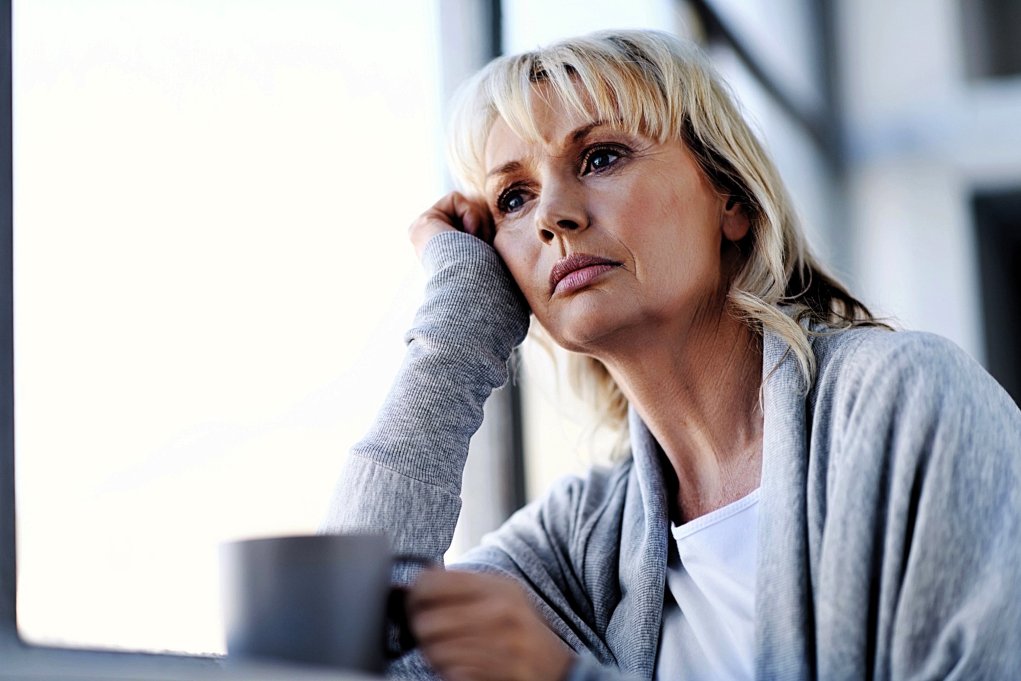 A woman with blonde hair and a thoughtful expression leans her head on her hand while looking out of a window. She is wearing a gray sweater and holding a mug, sitting in a brightly lit room.