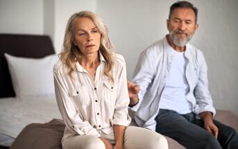 An older couple sits on a bed. The woman in the foreground looks upset and gazes away, while the man in the background reaches out towards her, appearing concerned. The room is softly lit and minimalistic in style.