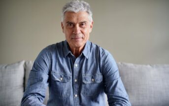 A man with short gray hair is sitting on a couch. He is wearing a denim button-up shirt and smiling slightly. The background is plain and neutral in color.