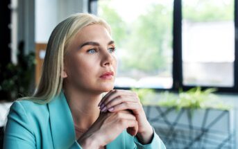 A person with long blonde hair sits indoors, looking thoughtfully out of a window. They are wearing a light blue blazer and have their hands clasped under their chin. The background features a decorative wall and green plants.