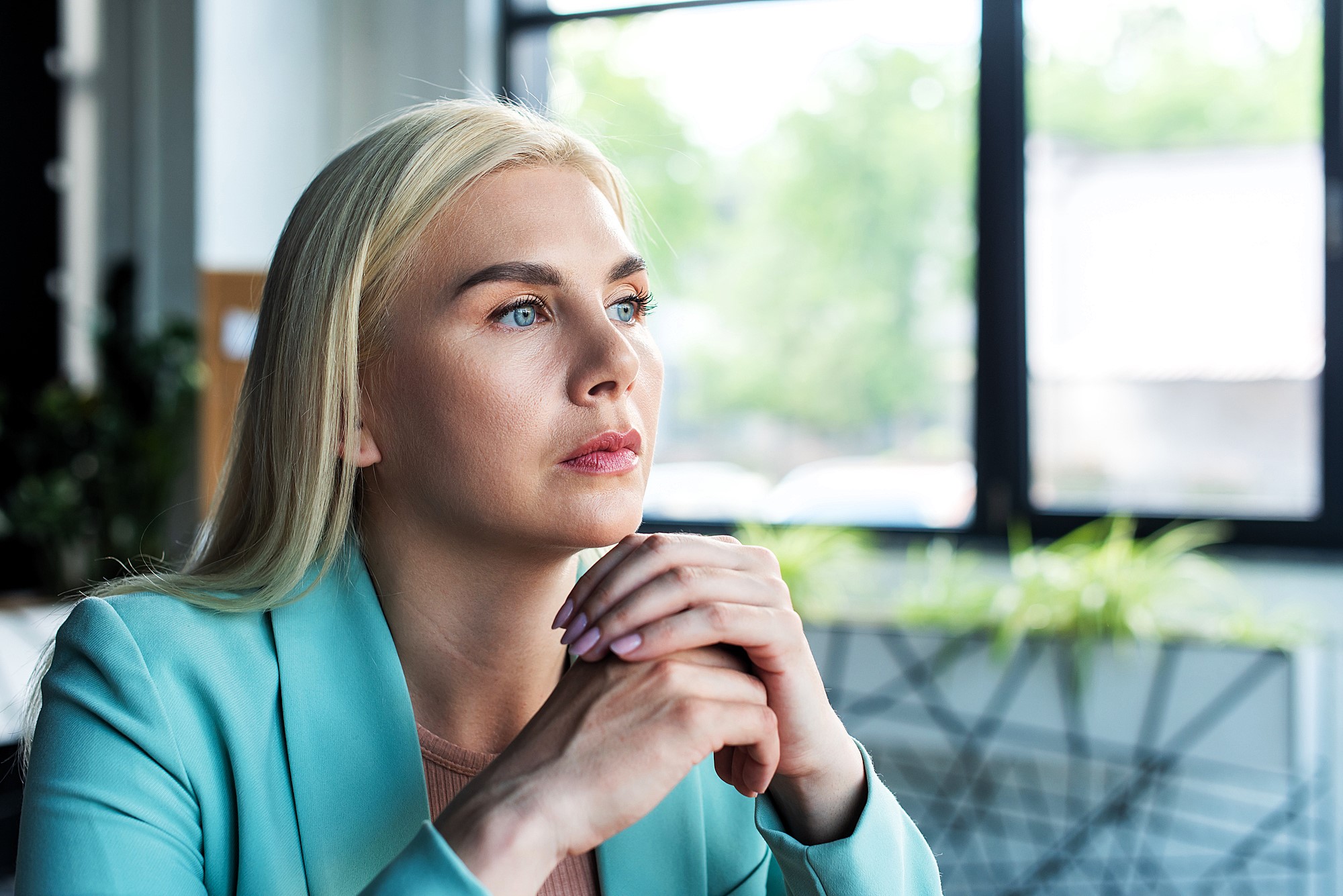 A person with long blonde hair sits indoors, looking thoughtfully out of a window. They are wearing a light blue blazer and have their hands clasped under their chin. The background features a decorative wall and green plants.