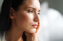 Close-up of a pensive woman resting her chin on her hand. She gazes thoughtfully into the distance with soft lighting highlighting her features. The background is blurred, creating a serene atmosphere.