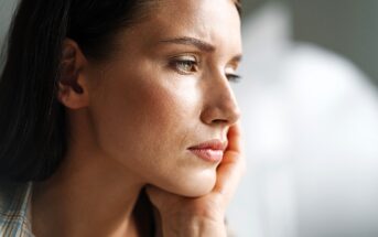 Close-up of a pensive woman resting her chin on her hand. She gazes thoughtfully into the distance with soft lighting highlighting her features. The background is blurred, creating a serene atmosphere.