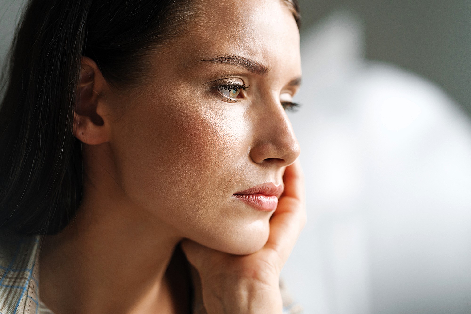 Close-up of a pensive woman resting her chin on her hand. She gazes thoughtfully into the distance with soft lighting highlighting her features. The background is blurred, creating a serene atmosphere.