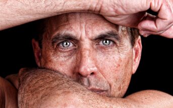 A close-up of a person with intense eyes, resting their head on their arm. The image focuses on the facial expression, highlighting the eyes and skin texture against a dark background.