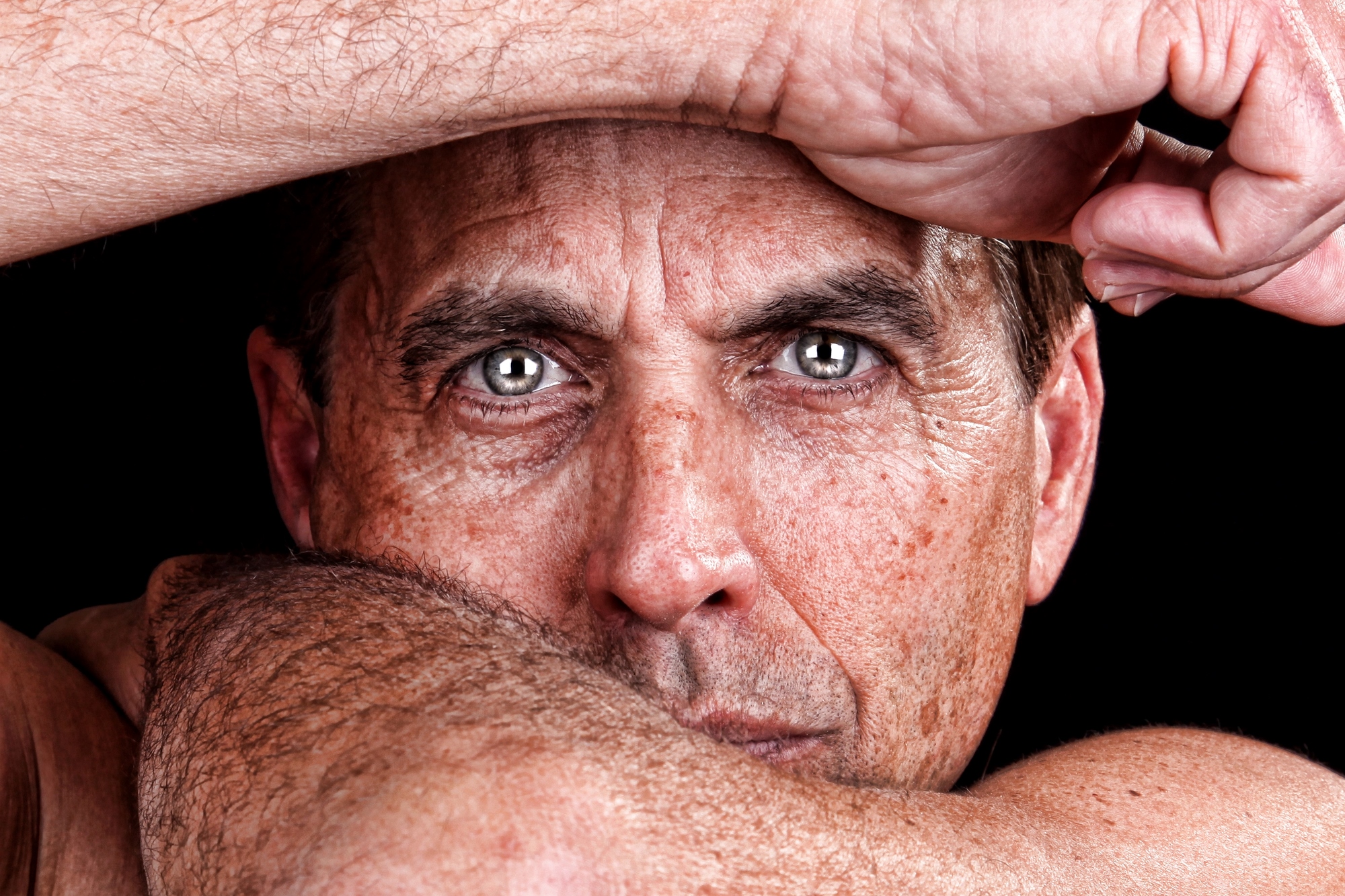 A close-up of a person with intense eyes, resting their head on their arm. The image focuses on the facial expression, highlighting the eyes and skin texture against a dark background.