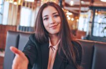 A woman with long brown hair sits in a restaurant, smiling and reaching forward as if to shake hands. She is wearing a black blazer over a light-colored blouse. The background is softly lit with warm lights.