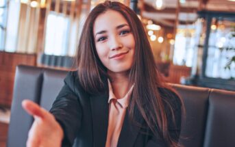 A woman with long brown hair sits in a restaurant, smiling and reaching forward as if to shake hands. She is wearing a black blazer over a light-colored blouse. The background is softly lit with warm lights.