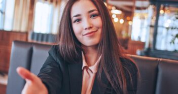 A woman with long brown hair sits in a restaurant, smiling and reaching forward as if to shake hands. She is wearing a black blazer over a light-colored blouse. The background is softly lit with warm lights.