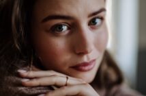 Close-up of a person with light blue-green eyes gazing directly at the camera. Their hands, with a simple ring, rest near their face, and they have light makeup with a soft expression. The background is blurred.