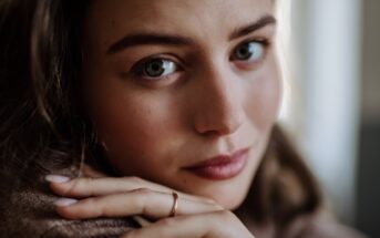 Close-up of a person with light blue-green eyes gazing directly at the camera. Their hands, with a simple ring, rest near their face, and they have light makeup with a soft expression. The background is blurred.