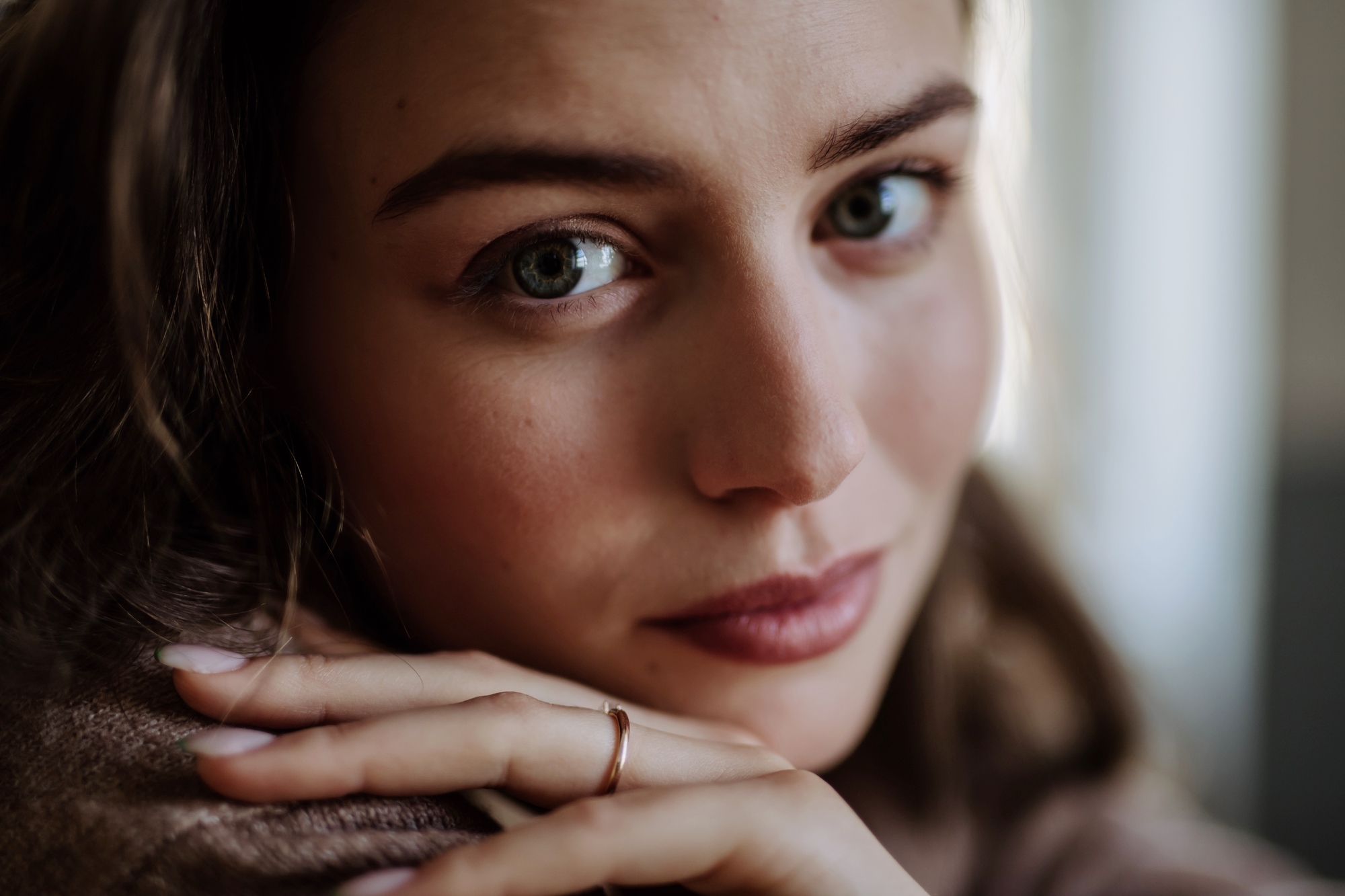 Close-up of a person with light blue-green eyes gazing directly at the camera. Their hands, with a simple ring, rest near their face, and they have light makeup with a soft expression. The background is blurred.