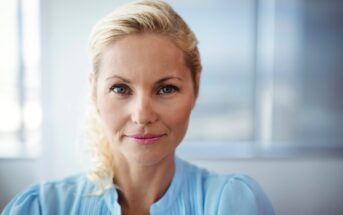 A woman with blonde hair wearing a light blue shirt smiles softly at the camera. She is indoors with a blurred background.