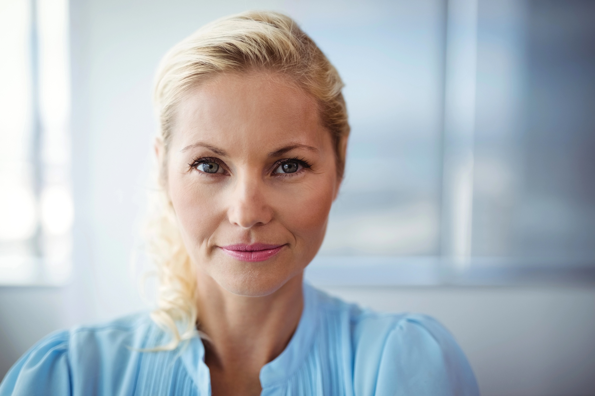 A woman with blonde hair wearing a light blue shirt smiles softly at the camera. She is indoors with a blurred background.