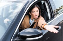 A person with long dark hair, wearing a tan coat and light blue scarf, sits in a car on the driver's side, gesturing outside the window with an expressive face. The car is black and the side mirror is visible.