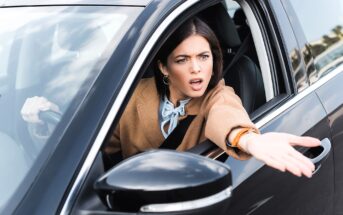 A person with long dark hair, wearing a tan coat and light blue scarf, sits in a car on the driver's side, gesturing outside the window with an expressive face. The car is black and the side mirror is visible.