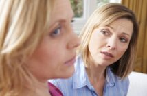 Two women indoors, one in focus looking concerned and the other slightly blurred, facing away. The background is softly lit, suggesting a home or casual setting.