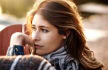 A woman with long hair gazes thoughtfully into the distance. She is wearing a patterned scarf and a jacket, with her chin resting on her hand. The background is softly blurred, suggesting an outdoor setting. The lighting is warm and gentle.