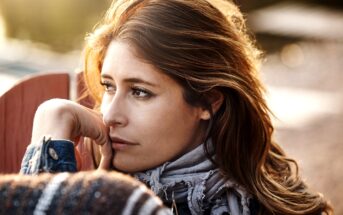 A woman with long hair gazes thoughtfully into the distance. She is wearing a patterned scarf and a jacket, with her chin resting on her hand. The background is softly blurred, suggesting an outdoor setting. The lighting is warm and gentle.