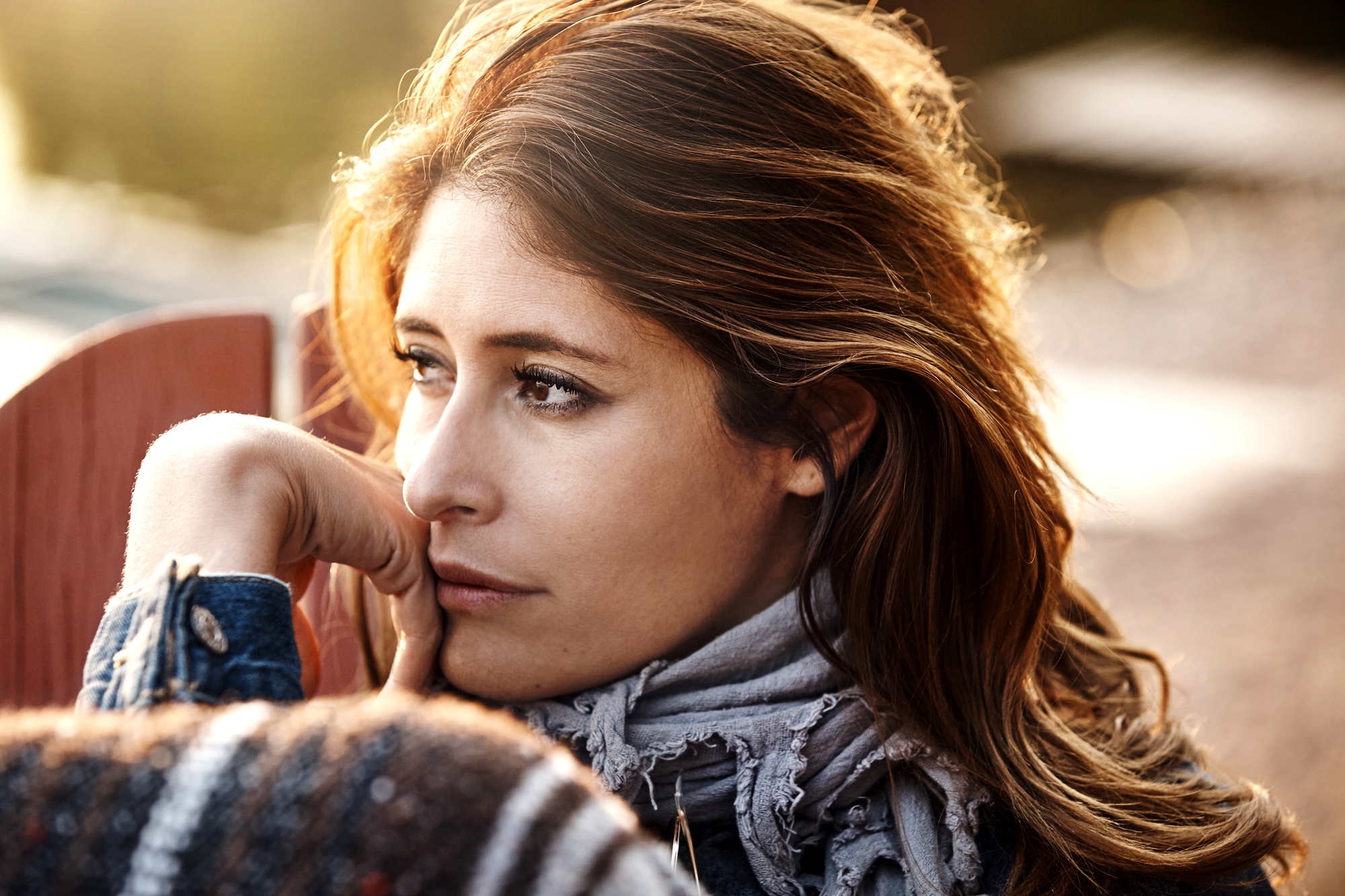 A woman with long hair gazes thoughtfully into the distance. She is wearing a patterned scarf and a jacket, with her chin resting on her hand. The background is softly blurred, suggesting an outdoor setting. The lighting is warm and gentle.