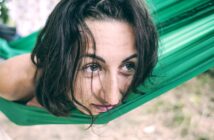 A person with wavy brown hair is resting in a green hammock, gazing thoughtfully into the distance. The background is blurred, suggesting an outdoor setting with greenery.