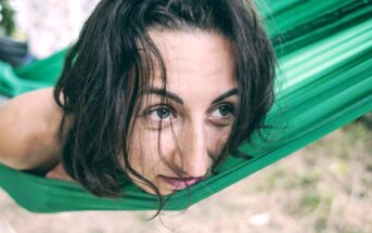 A person with wavy brown hair is resting in a green hammock, gazing thoughtfully into the distance. The background is blurred, suggesting an outdoor setting with greenery.